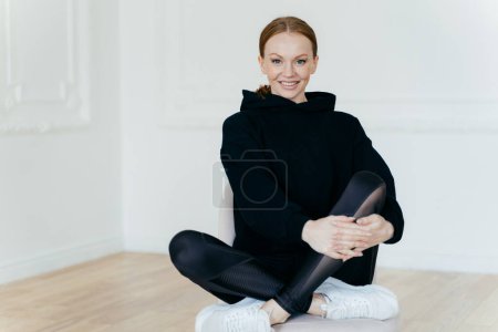 Photo for Positive European sporty woman in good mood has make up, sits crossed legs on comfortable chair, has joyful expression, wears black sweatshirt and leggings, poses indoor, has pleasant smile on face. - Royalty Free Image