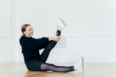 Photo for Athletic woman stretches leg, has satisfied facial expression, wears black clothes, white sportshoes, sits on floor, does pilates exercises, has joyful look, exercises acitively. Sport concept - Royalty Free Image