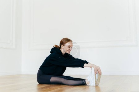 Photo for Indoor shot of smiling redhead woman stretches legs, focused into distance, touches feet, wears white sportshoes, has gymnastic exercises, prepares for cardio training. Healthy lifestyle concept - Royalty Free Image