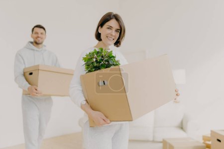 Photo for Happy young couple enter own modern house, buy real estate, carry cardboard boxes with indoor plant and other personal stuff, buy new house for living, have moving day. People and property concept - Royalty Free Image
