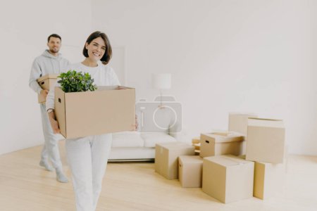 Photo for Happy woman and her husband carry boxes with personal belongings, being busy during relocation in other place for living, enter new home, move together in big house, unpack household things. - Royalty Free Image