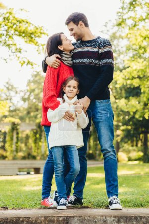 Photo for Young happy family of three stand together outdoors, embrace, have good relationships. Couple in love look at each other, hug their little daughter. Happiness and harmony in family life. - Royalty Free Image
