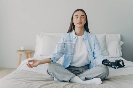 Disabled woman meditates in lotus pose on bed. Caucasian girl with sensored bionic prosthesis. Eyes closed, myoelectric limb. Stress relief.