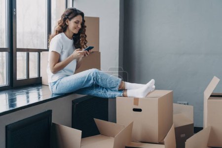 Photo for Girl homeowner tenant chooses moving company via smartphone app. Waiting with cardboard boxes on windowsill for house relocation - Royalty Free Image