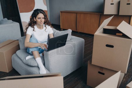 Photo for Dreaming of a new home, modern girl works on laptop amidst cardboard boxes. Pensive woman shops online, plans apartment interior. Relocation and house repair aspirations. - Royalty Free Image