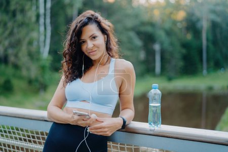 Photo for Athletic brunette in top and leggings on bridge, listening to music, using phone, staying hydrated. Active lifestyle. - Royalty Free Image