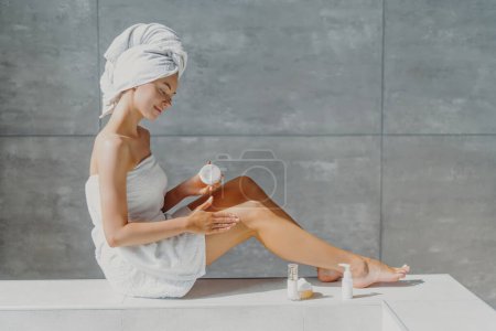 Pretty woman applies moisturizing cream, slender legs. Beauty routine in bathroom. Softness after lotion.