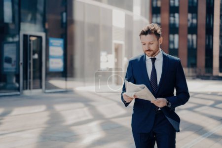 Photo for Successful businessman reads newspaper, walks by glass office building in financial district on sunny morning. - Royalty Free Image