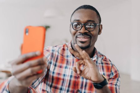 Smiling African-American man in glasses shows okay sign, communicates on video call, holds smartphone.