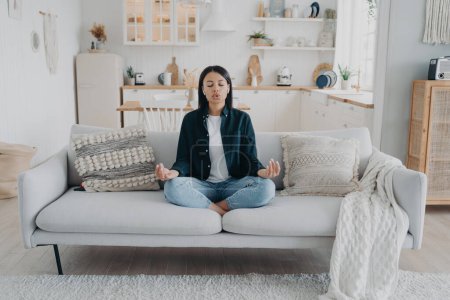 Photo for Serene woman practice yoga with mudra gesture, breathing sitting on sofa in living room. Young female meditate relieve stress, relaxing at home. Healthy lifestyle, emotion management. - Royalty Free Image