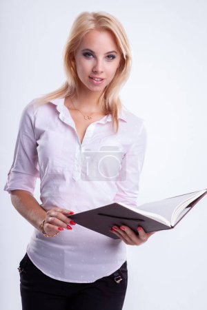 Photo for Portrait of a  young adult attractive blonde businesswoman in pink shirt reading notebook on light backround - Royalty Free Image