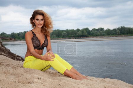 Foto de Retrato de sensualidad atractiva y bonita mujer sexy rubia en pantalones amarillos y sujetador negro posando en la playa - Imagen libre de derechos