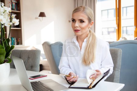Téléchargez les photos : Portrait de jeune adulte jolie femme d'affaires blonde en chemise blanche travaillant au bureau à domicile - en image libre de droit