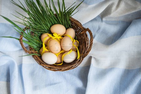 Téléchargez les photos : Beaux œufs de Pâques peints, un œuf avec un visage mignon, les œufs se trouvent dans un panier en bois avec de l'herbe verte. Carte postale de Pâques - en image libre de droit