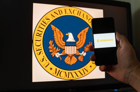 Téléchargez les photos : Kiev, Ukraine - 06 Juin 2023 : Homme tient un smartphone avec échange cryptographique Binance à l'écran sur le fond du moniteur avec le logo SEC. S.E.C. poursuivi sur Binance par blanchiment d'argent en BNB - en image libre de droit