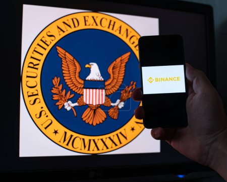 Téléchargez les photos : Kiev, Ukraine - 06 Juin 2023 : Homme tient un smartphone avec échange cryptographique Binance à l'écran sur le fond du moniteur avec le logo SEC. S.E.C. poursuivi sur Binance par blanchiment d'argent en BNB - en image libre de droit