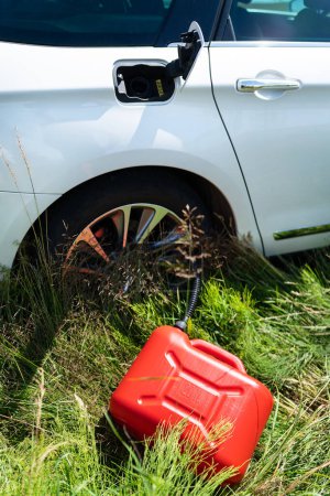 Foto de Bote de plástico rojo entre la hierba verde cerca del coche blanco. Problemas en la carretera con el combustible para el coche. Foto vertical - Imagen libre de derechos