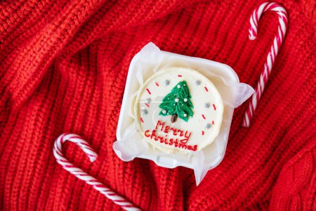 A festive Christmas cupcake with a white frosting and a green tree on top, on a red knitted background with candy canes. Merry Christmas lettering