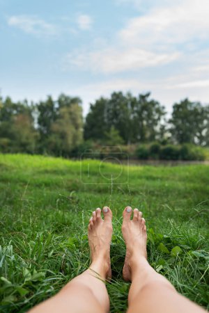 Photo for The image shows a pair of bare feet lying on a grassy field with a lake and a green park in the background. Toes point upward. The image is peaceful and calm - Royalty Free Image