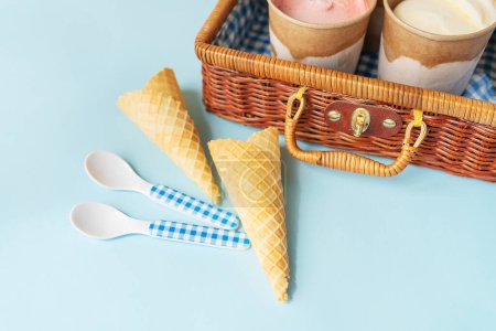 Téléchargez les photos : L'image montre des cônes et des tasses de crème glacée dans un panier de pique-nique en osier sur un fond bleu. L'image est idéale pour les thèmes d'été, pique-nique et dessert - en image libre de droit