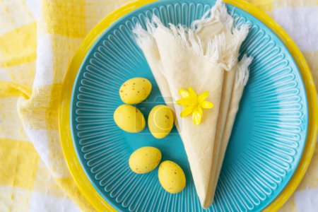 This vibrant Easter table setting features a blue plate adorned with yellow eggs and a delicate flower. The checkered tablecloth adds a festive touch
