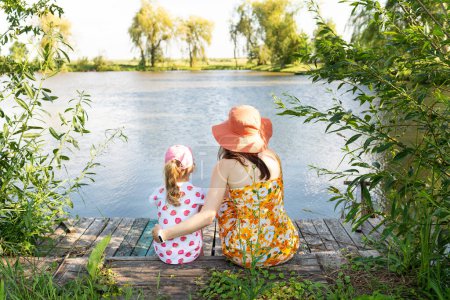 Mother and daughter sit by a serene lake surrounded by lush greenery, evoking a sense of peace and connection with nature
