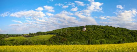 Téléchargez les photos : Champ de colza jaune printemps vue champs, ciel bleu avec nuages dans le panorama de la lumière du soleil. Tour Pyatnychany (structure de défense, 15ème siècle) sur la pente de la colline lointaine. - en image libre de droit