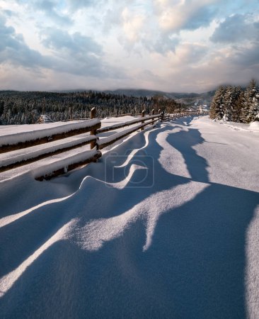 Foto de Pintorescas sombras ondeadas en la nieve de la cerca de madera. Montaña alpina invierno aldea periferia, sendero nevado, bosque de abeto. Imagen de alta resolución con gran profundidad de campo. - Imagen libre de derechos