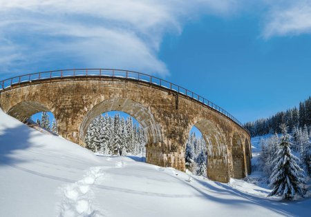 Téléchargez les photos : Viaduc en pierre (pont en arc) sur chemin de fer à travers la forêt de sapins neigeux de montagne. Dérives de neige sur le bord de la route et givre sur les arbres et les fils électriques. - en image libre de droit
