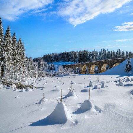 Foto de Viaducto de piedra (puente de arco) en el ferrocarril a través del bosque de abeto nevado de montaña. La nieve se desplaza por el camino y acumula escarcha en los árboles y cables eléctricos. - Imagen libre de derechos