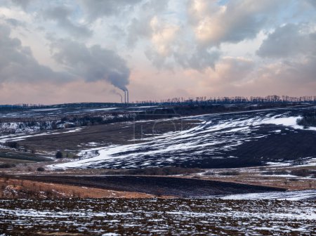 Foto de Atardecer tranquilo en la campiña ucraniana. Tierras cultivables cubiertas por la última nieve, principios de primavera Chimeneas industriales de Burshtyn TPP en el horizonte. - Imagen libre de derechos