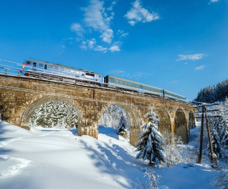Foto de Viaducto de piedra (puente de arco) en el ferrocarril a través del bosque de abeto nevado de montaña. La nieve se desplaza por el camino y acumula escarcha en los árboles y cables eléctricos. - Imagen libre de derechos