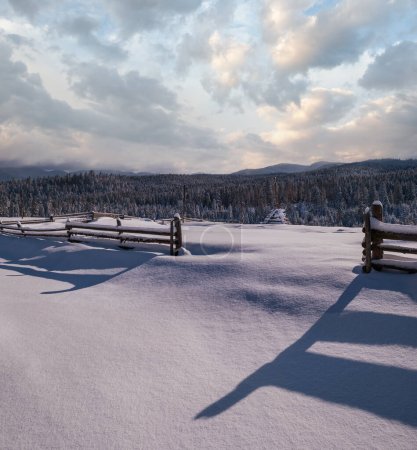 Foto de Pintorescas sombras en la nieve de la cerca de madera. Montaña alpina invierno aldea periferia, sendero nevado, bosque de abeto en colinas lejanas brumosas y nubladas. - Imagen libre de derechos