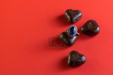 Photo for Heart shaped dark chocolates with a blue sapphire ring on a red background for holidays - Royalty Free Image