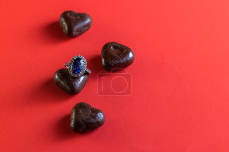 Foto de Heart shaped dark chocolates with a blue sapphire ring on a red background for holidays - Imagen libre de derechos