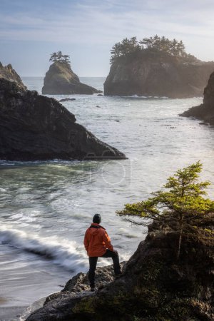 Foto de Hombre sentado en una roca disfrutando de la increíble vista del mar y las pilas de mar en este hermoso lugar en la costa de Oregon - Imagen libre de derechos