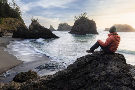 Téléchargez les photos : Homme assis sur un rocher profitant de la vue imprenable sur la mer et les piles de mer dans ce bel endroit sur la côte de l'Oregon - en image libre de droit
