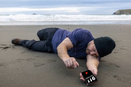 Téléchargez les photos : Man trying to call 911 after collapsing on the beach and holding his chest and showing signs of pain - en image libre de droit