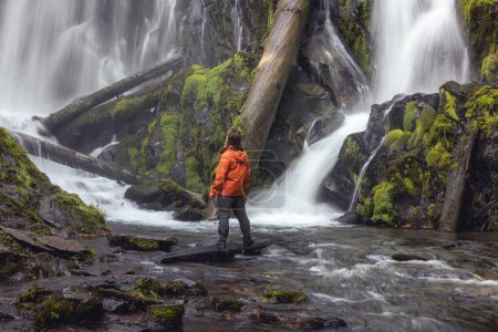 Foto de Caminante con una chaqueta naranja de pie frente a una cascada grande y hermosa en el bosque de Oregon - Imagen libre de derechos