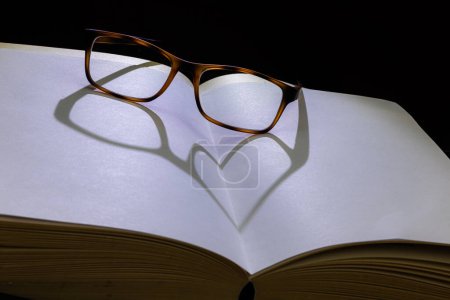 Téléchargez les photos : Ombre en forme de coeur sur un livre ouvert à partir d'une paire de lunettes rétro-éclairées - en image libre de droit