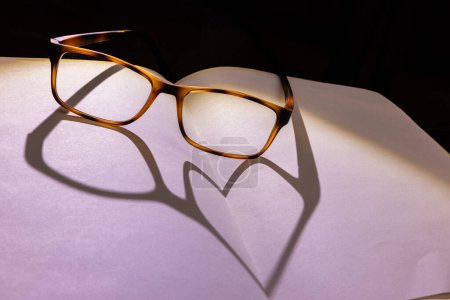 Téléchargez les photos : Ombre en forme de coeur sur un livre ouvert à partir d'une paire de lunettes rétro-éclairées - en image libre de droit