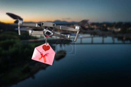 Photo for Drone carrying a pink letter with a red heart and bow flying over a lake and city in the background - Royalty Free Image