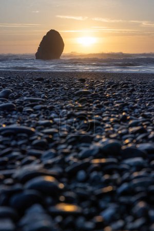 Ein felsiger Strand mit einem großen Felsen im Vordergrund und einem Sonnenuntergang im Hintergrund