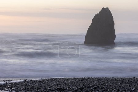 Foto de Una gran roca se encuentra en la playa, con el océano en el fondo. El agua está agitada y el cielo es una mezcla de rosa y azul. La escena es serena y pacífica - Imagen libre de derechos