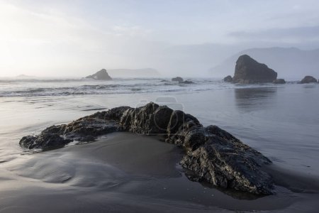 Une plage rocheuse avec un grand rocher au premier plan. Le ciel est nuageux et l'eau calme