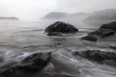 Téléchargez les photos : Un rivage rocheux avec un grand rocher au premier plan. L'eau est agitée et le ciel est couvert, créant une atmosphère lunatique et dramatique. Les rochers et l'eau semblent être en mouvement constant - en image libre de droit
