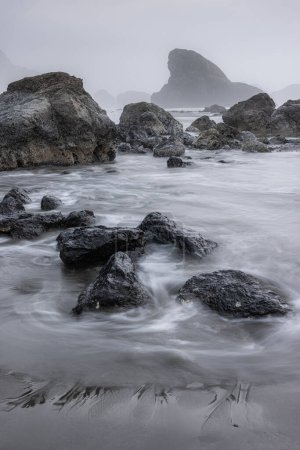 Foto de Una costa rocosa con una gran roca en la distancia. El agua está tranquila y el cielo está nublado. - Imagen libre de derechos