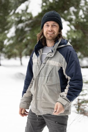 Téléchargez les photos : Un homme portant une veste bleue et grise et un chapeau noir sourit. Il est debout dans la neige avec des arbres en arrière-plan - en image libre de droit