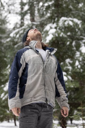 Téléchargez les photos : Un homme portant une veste grise et bleue est debout dans la neige. Il lève les yeux vers le ciel - en image libre de droit