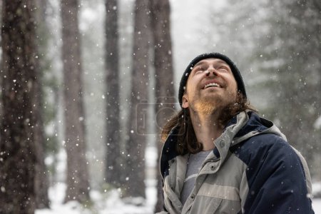 Téléchargez les photos : Un homme avec une barbe et un chapeau sourit dans la neige. Il porte une veste bleue et une chemise grise - en image libre de droit